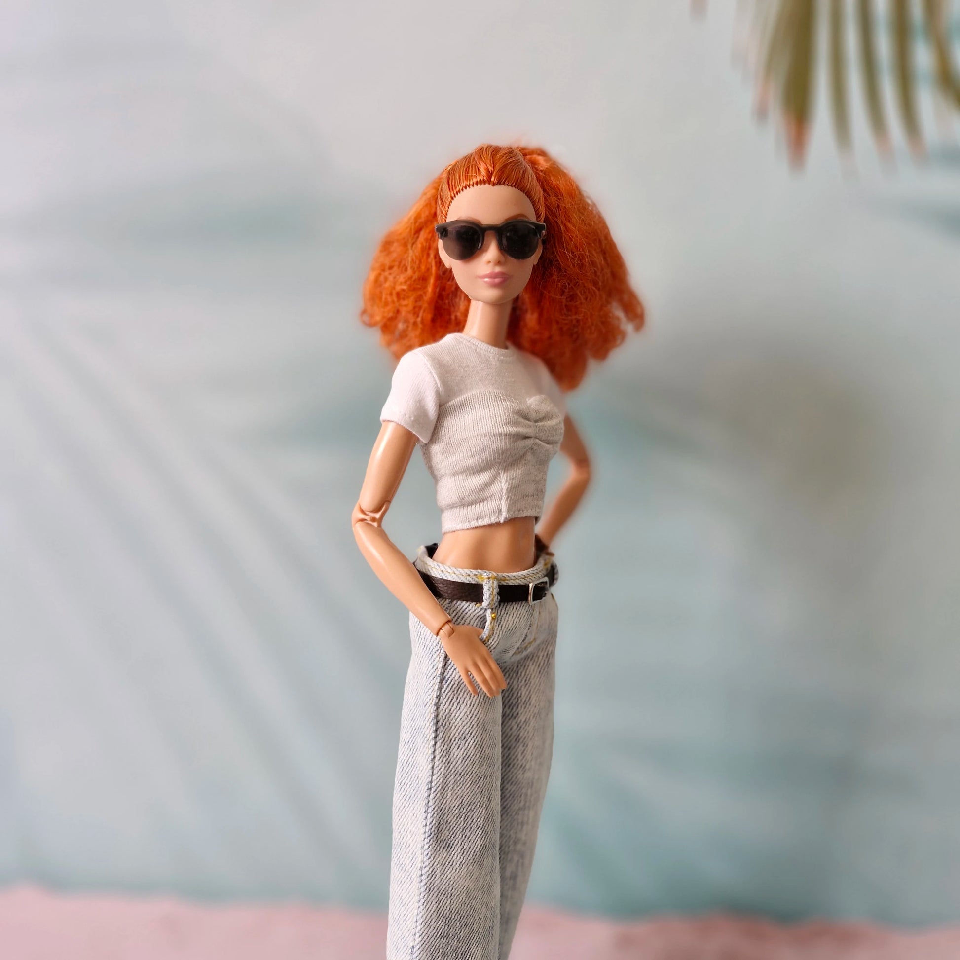 Barbie doll with red hair, sunglasses, white top, and gray pants against a blurred background
