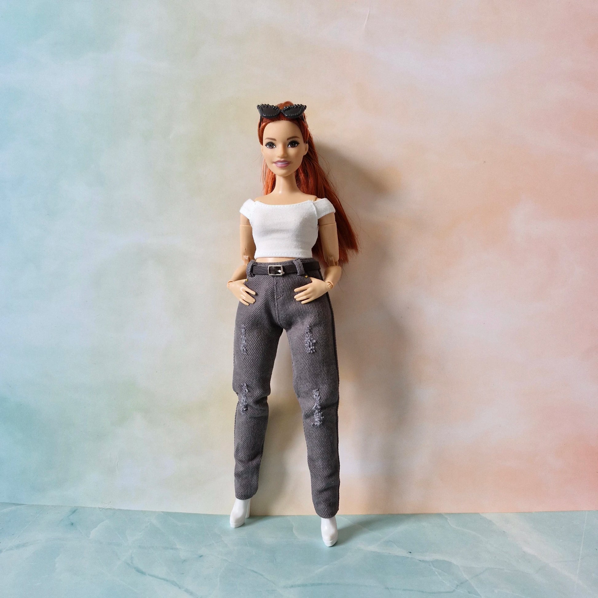 A red-haired Barbie doll models a modern outfit featuring slim-fit, distressed charcoal grey jeans with a black belt, a white ribbed crop top, and white boots, standing against a pastel background.