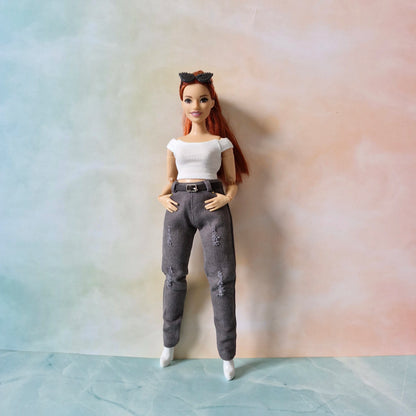 A red-haired Barbie doll models a modern outfit featuring slim-fit, distressed charcoal grey jeans with a black belt, a white ribbed crop top, and white boots, standing against a pastel background.