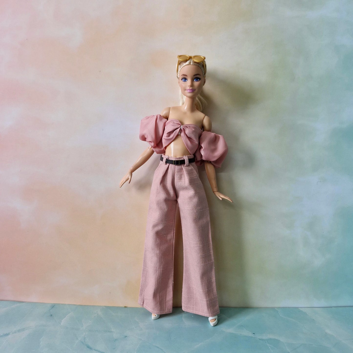 Barbie doll wearing a pink outfit with a bow top and wide-leg pants against a gradient background.