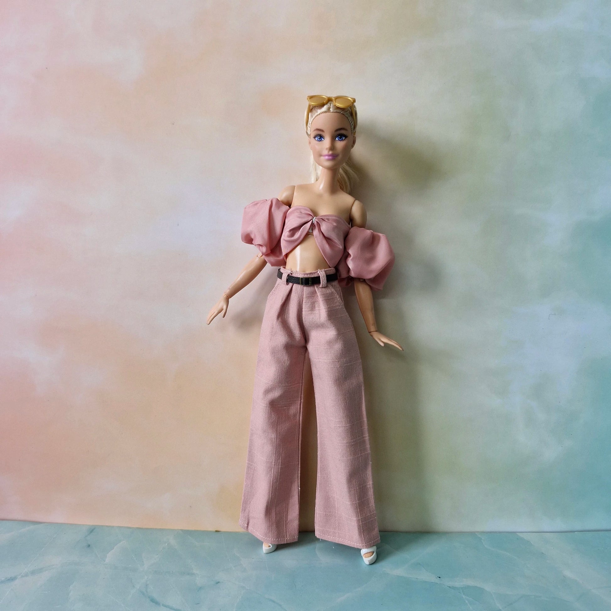 Barbie doll wearing a pink outfit with a bow top and wide-leg pants against a gradient background.
