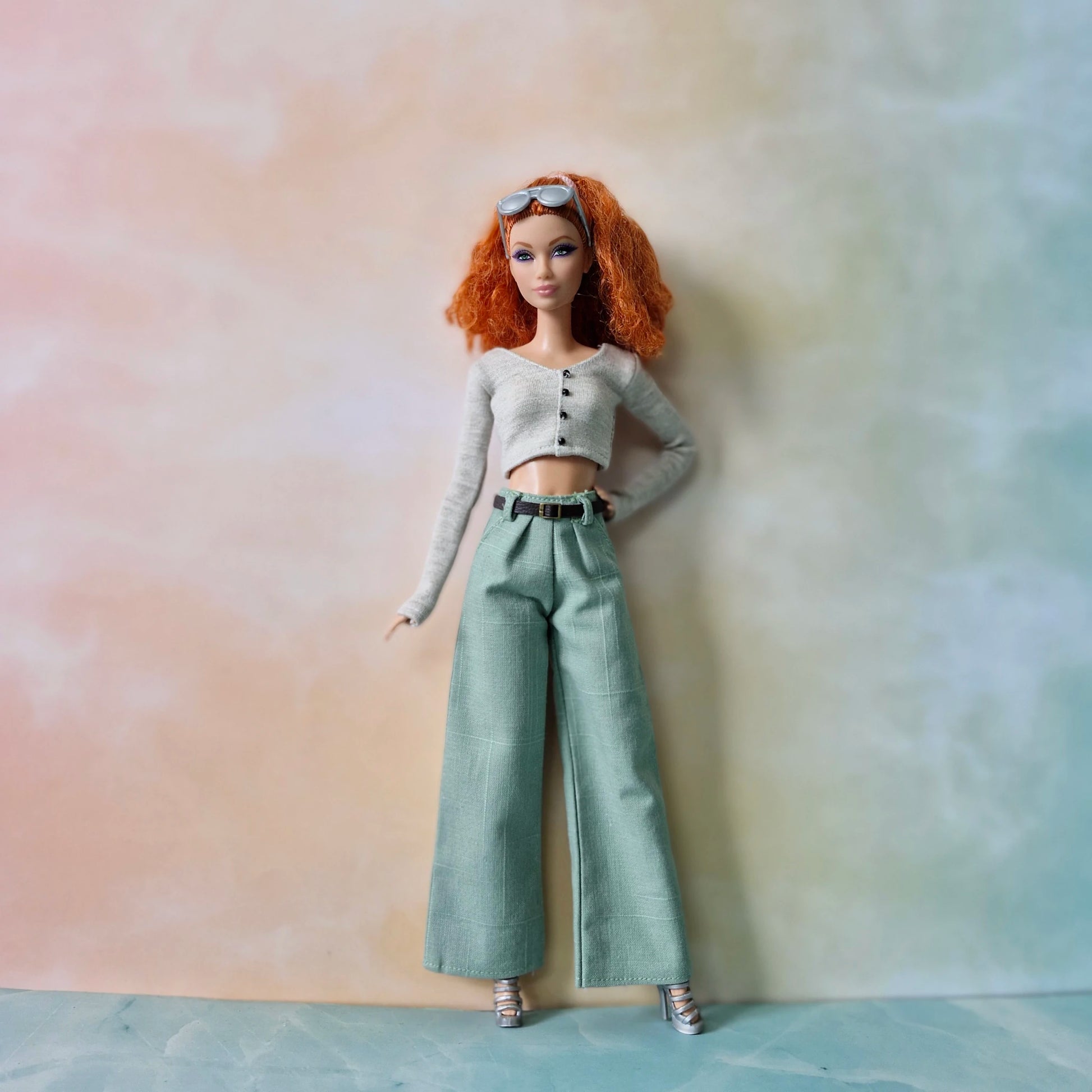 Barbie doll with red hair wearing a gray sweater and green pants on a neutral background