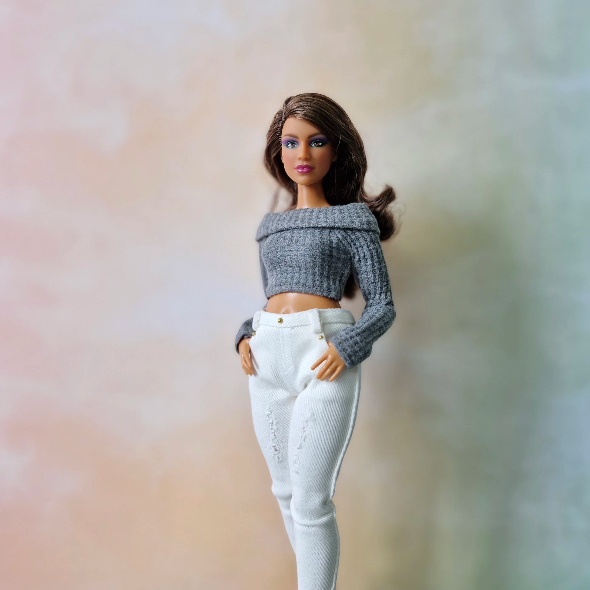 Barbie doll wearing a gray sweater and white pants against a blurred background