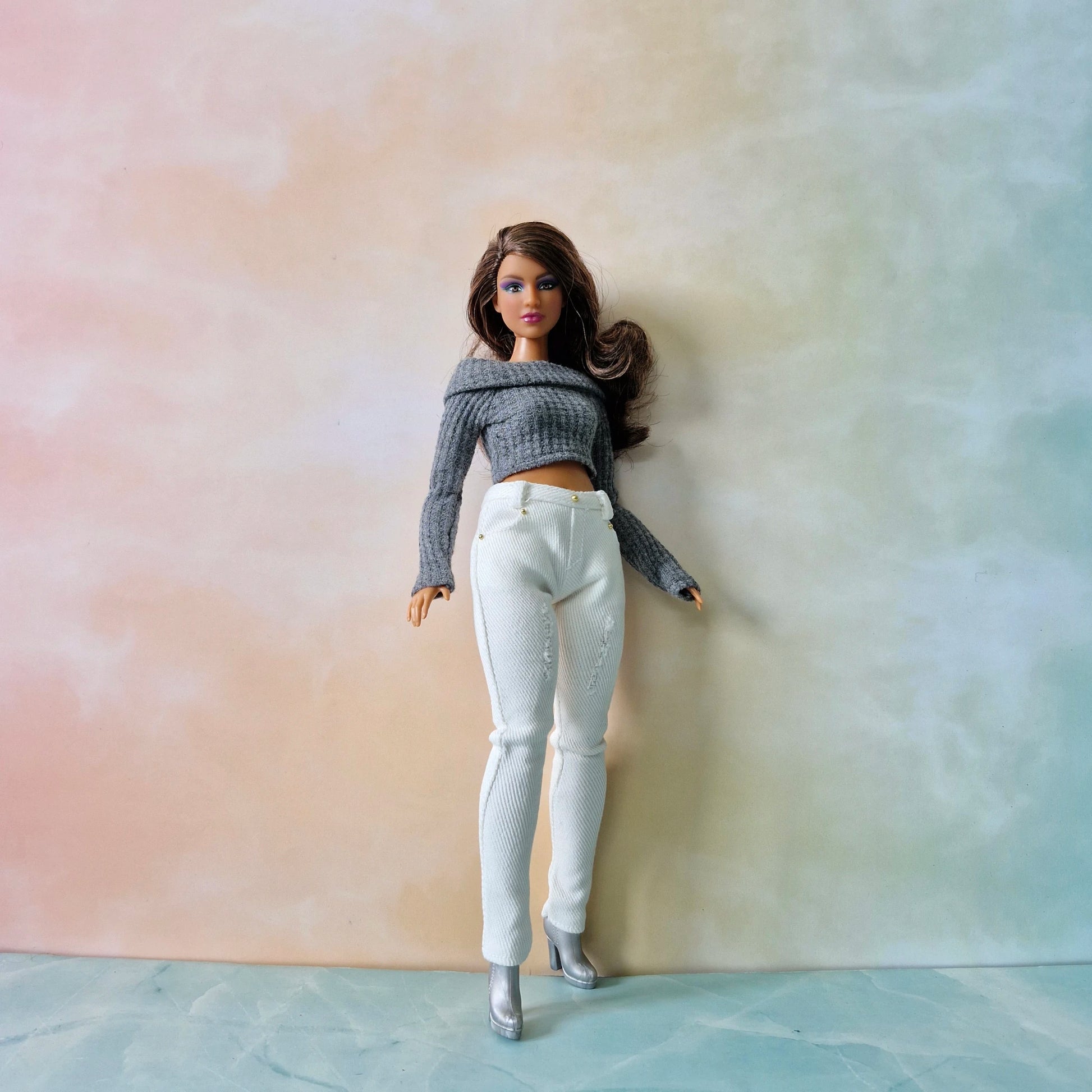 Barbie doll wearing a gray sweater and white pants against a pastel gradient background