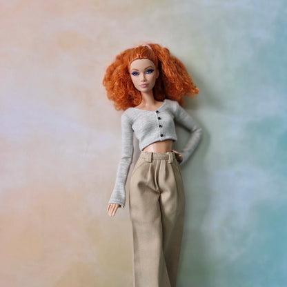Barbie doll wearing a gray sweater and beige pants against a gradient background