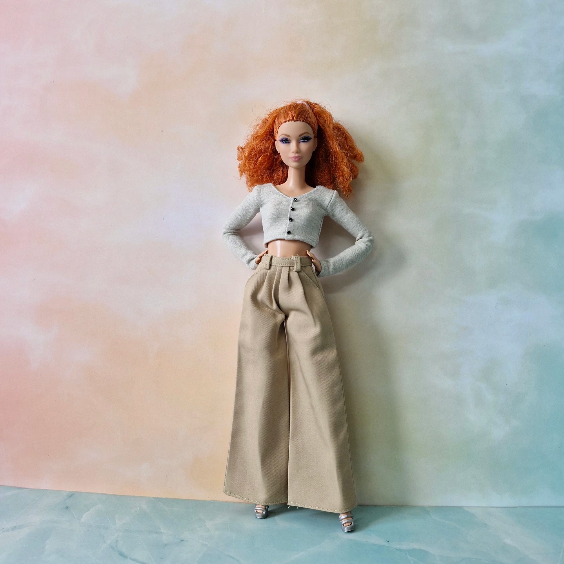 Barbie doll with red hair wearing a white top and beige pants against a pastel gradient background