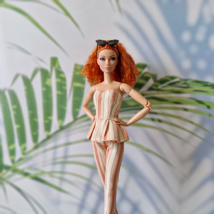 Barbie doll with red hair and a striped outfit standing against a leafy background