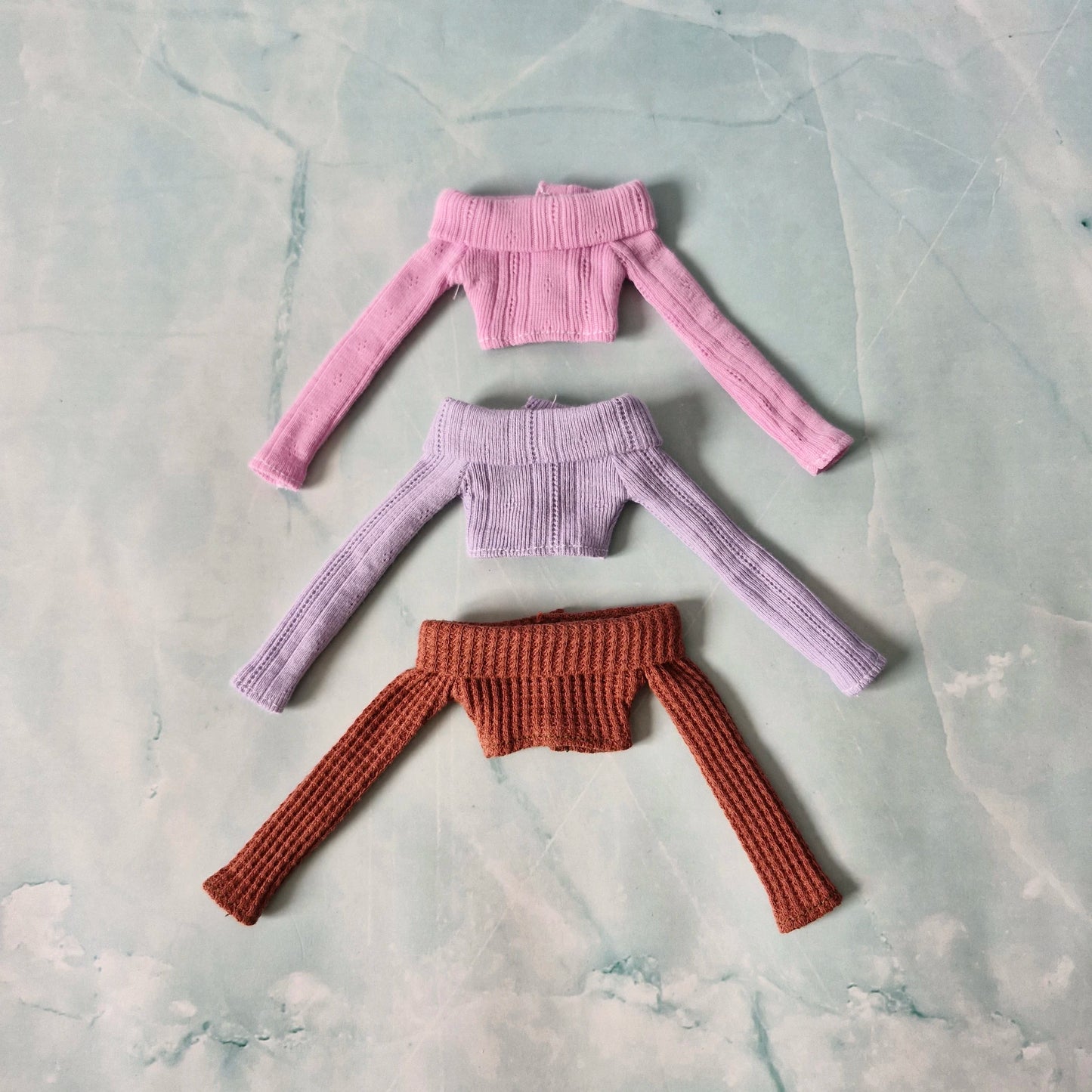 Three knitted tops in pink, purple, and brown on a textured gray surface