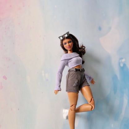 Barbie doll wearing a purple top and gray shorts against a pastel background