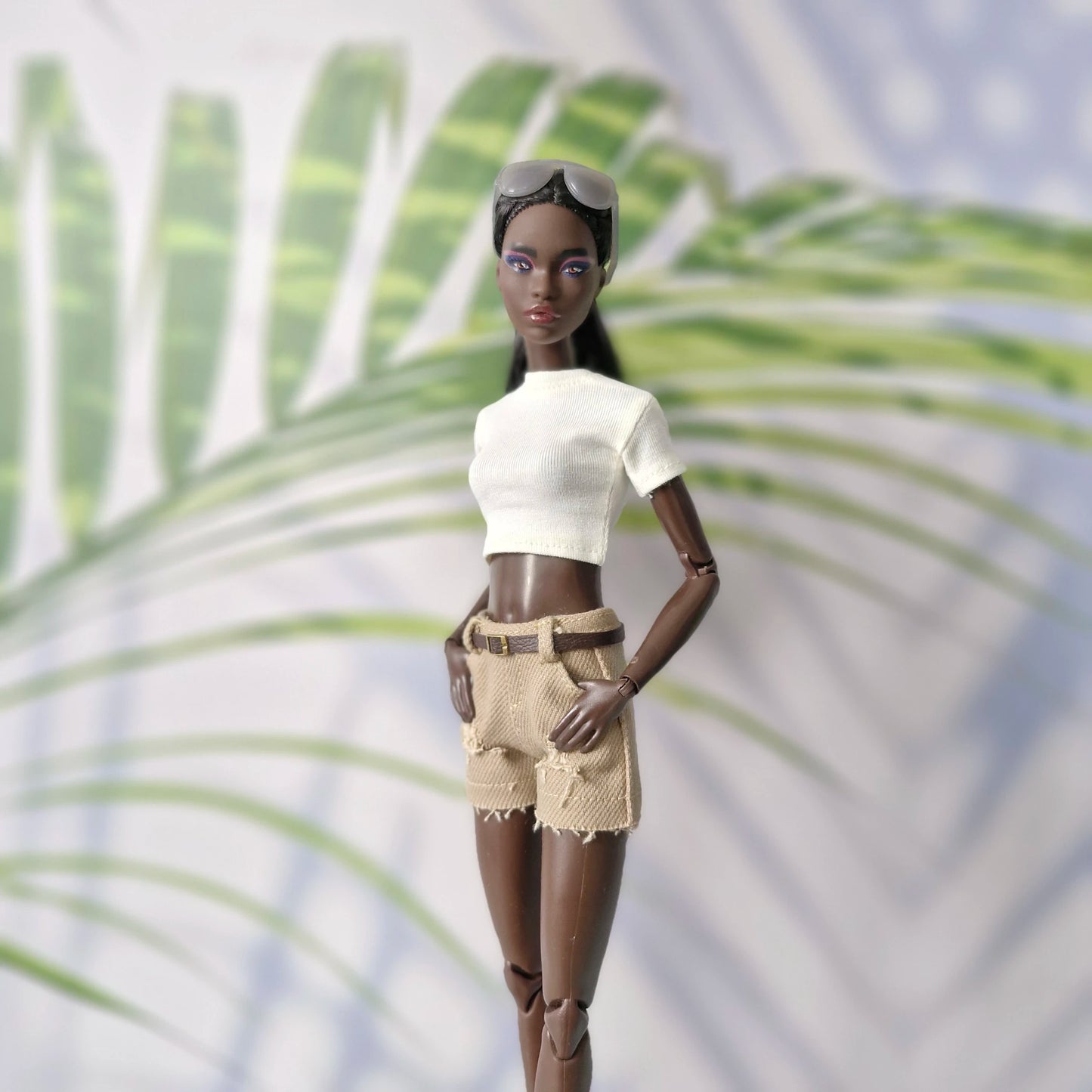 Barbie doll with brown skin wearing a white top and beige shorts against a palm leaf background
