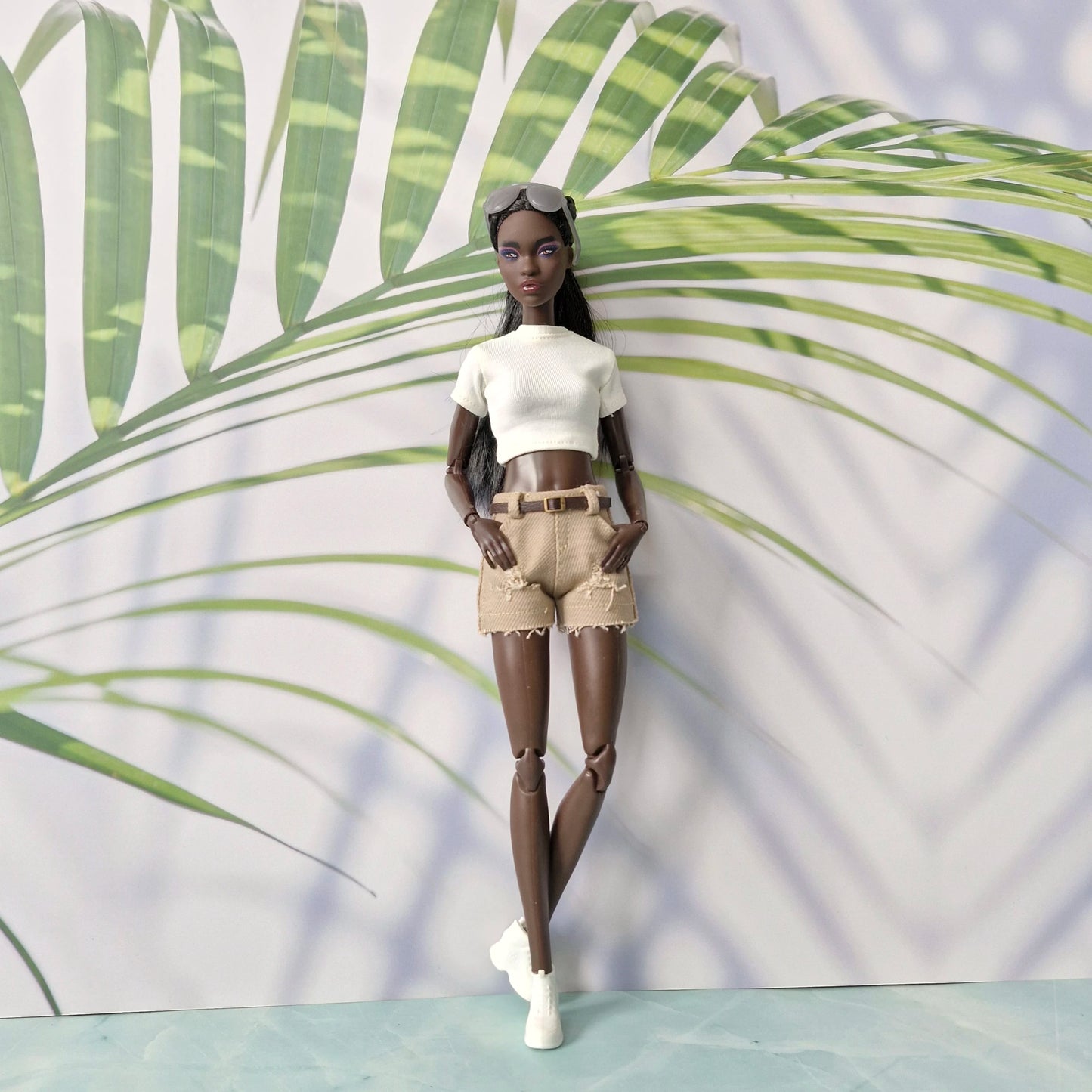 Doll wearing a white top and beige shorts against a leafy green background