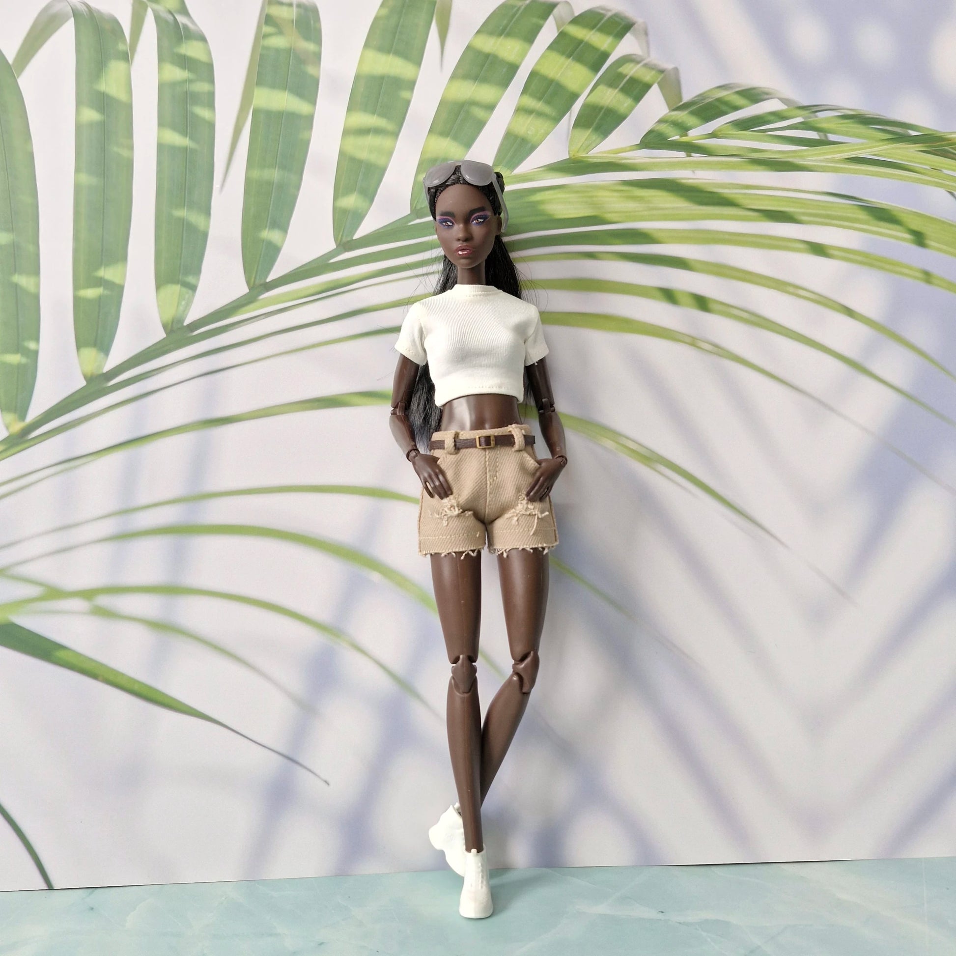 Doll wearing a white top and beige shorts against a leafy green background