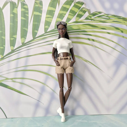 Doll wearing a white top and beige shorts against a leafy green background