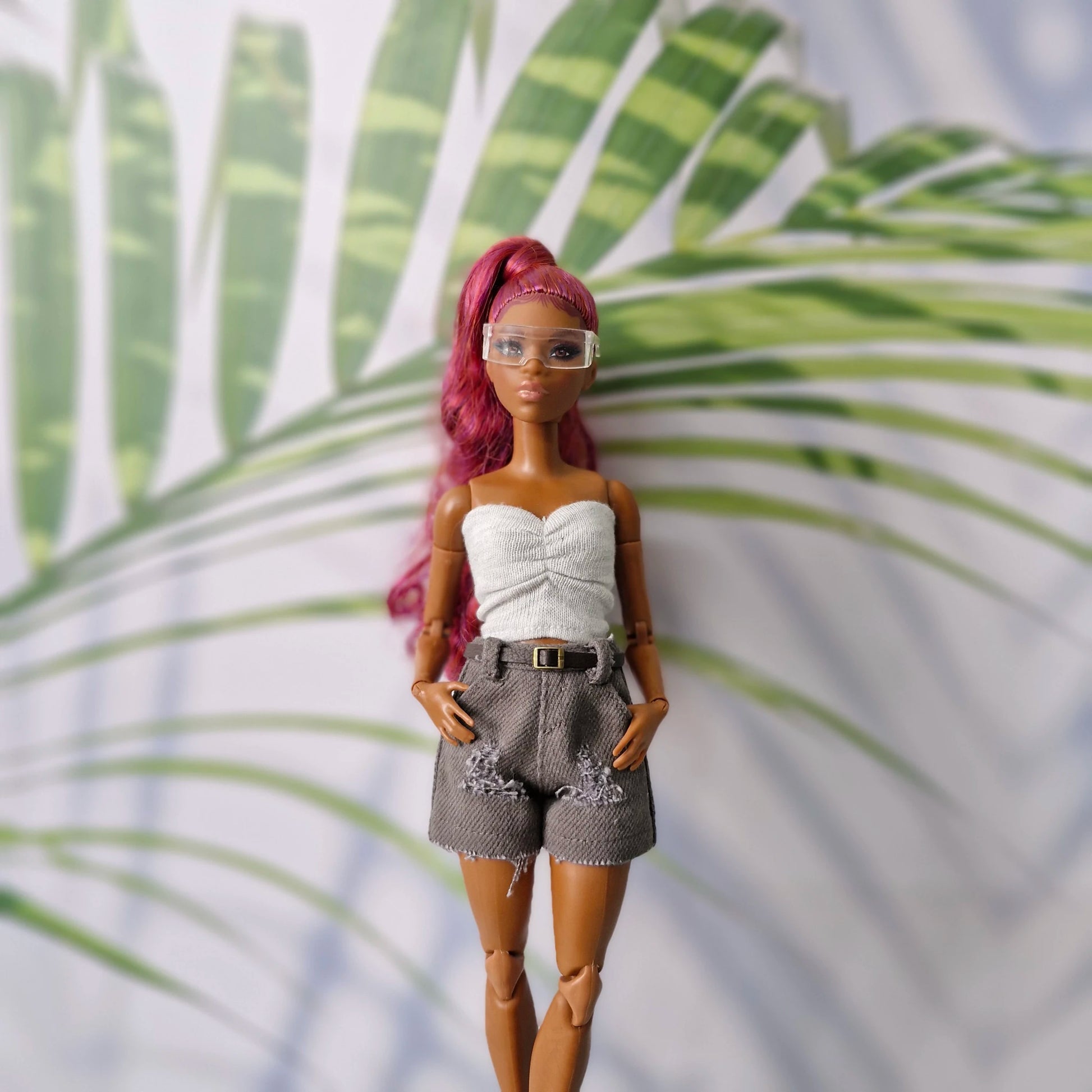 Barbie doll with pink hair and white top standing against a leafy green background