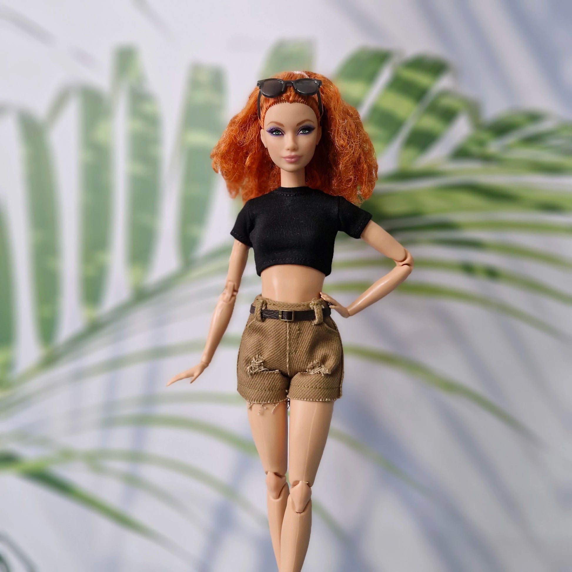 Barbie doll with red hair, black top, and brown shorts standing against a palm leaf background