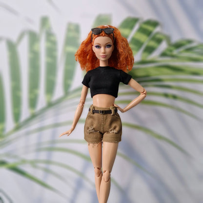 Barbie doll with red hair, black top, and brown shorts standing against a palm leaf background