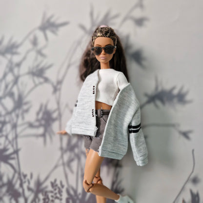 Barbie doll wearing a white top, gray cardigan, and sunglasses against a neutral background