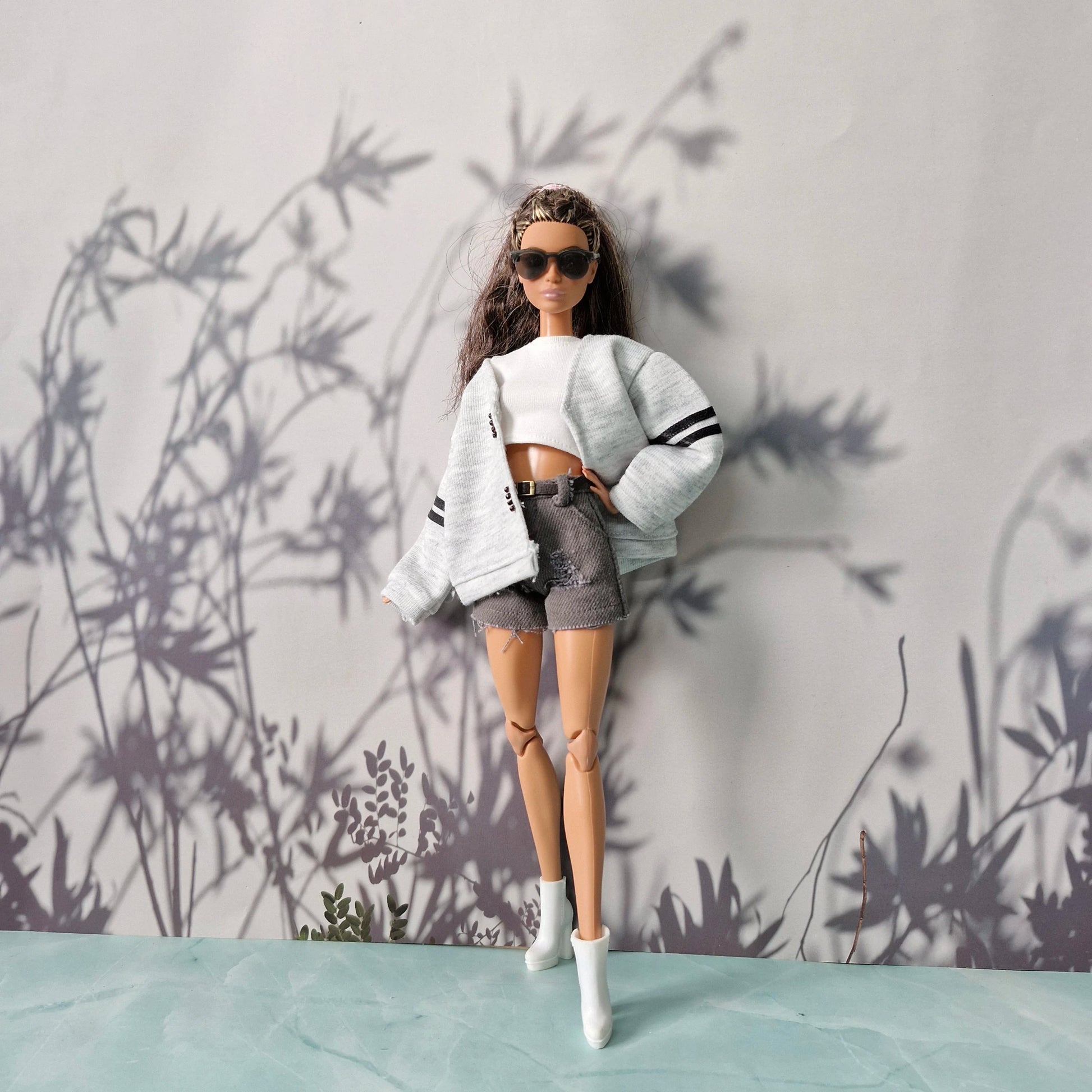 Barbie doll wearing a white sweater and gray shorts against a wall with plant shadows