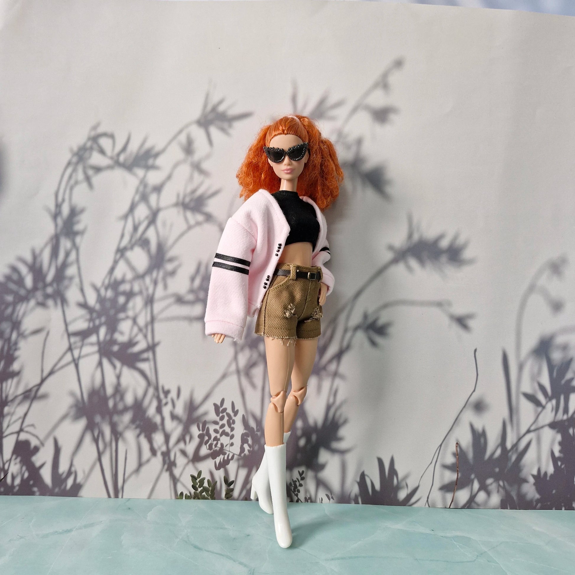 Barbie doll with red hair and sunglasses standing against a gray background with plant shadows.