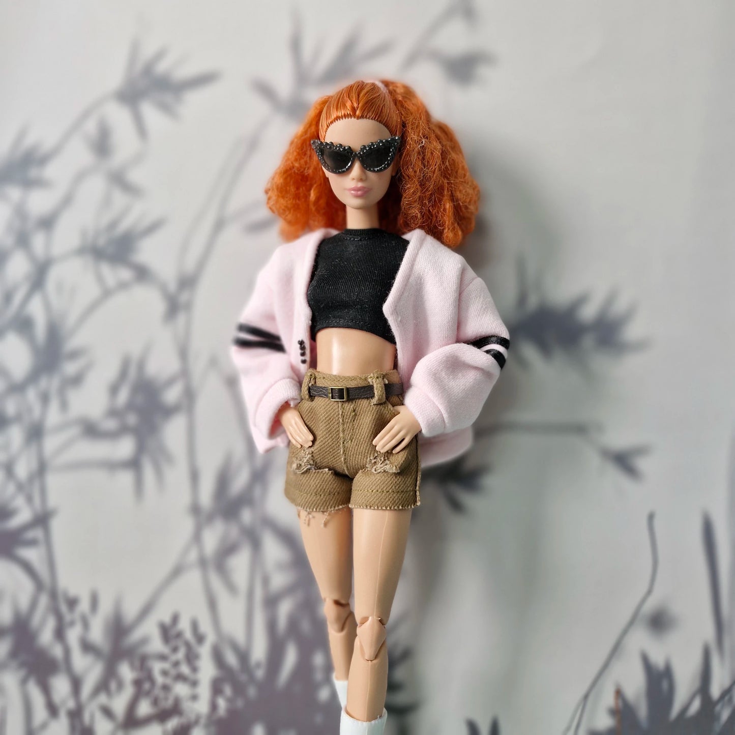 Barbie doll with red hair, sunglasses, pink cardigan, black top, and beige shorts against a gray background.