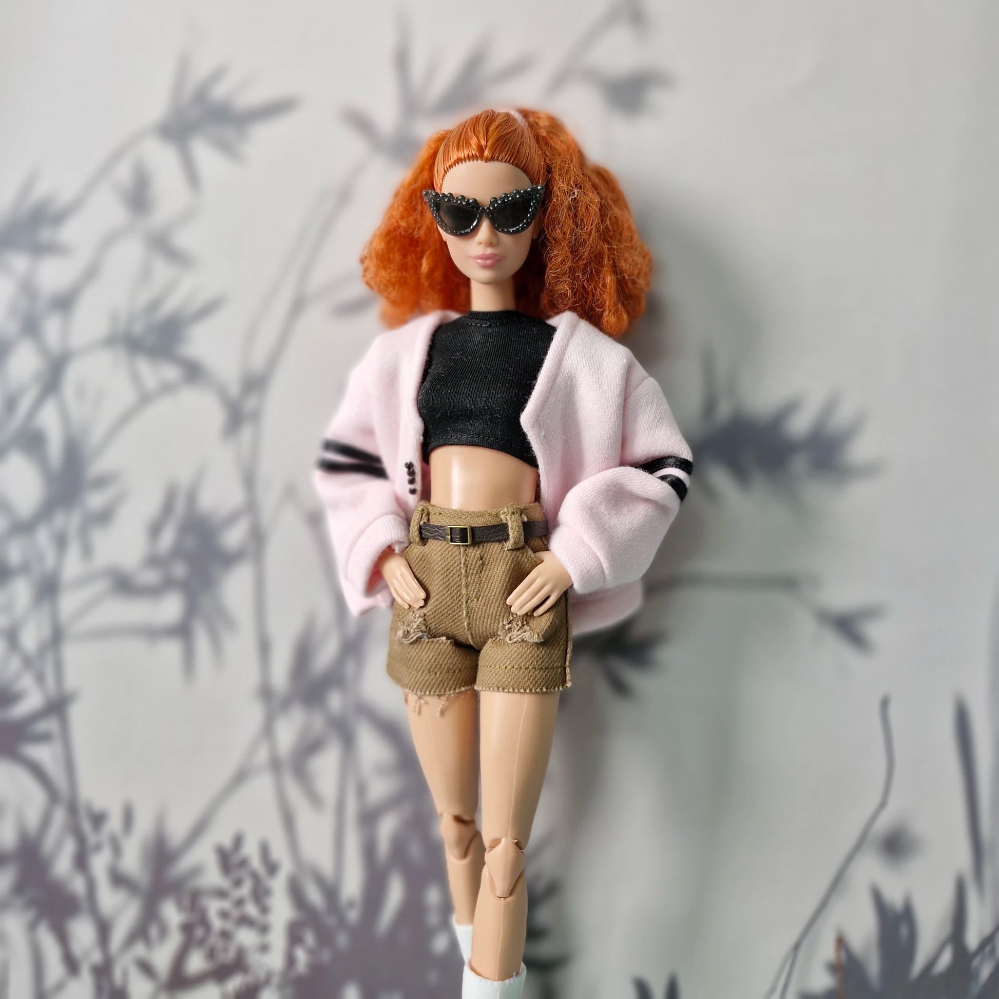 Barbie doll with red hair, sunglasses, pink cardigan, black top, and beige shorts against a gray background.