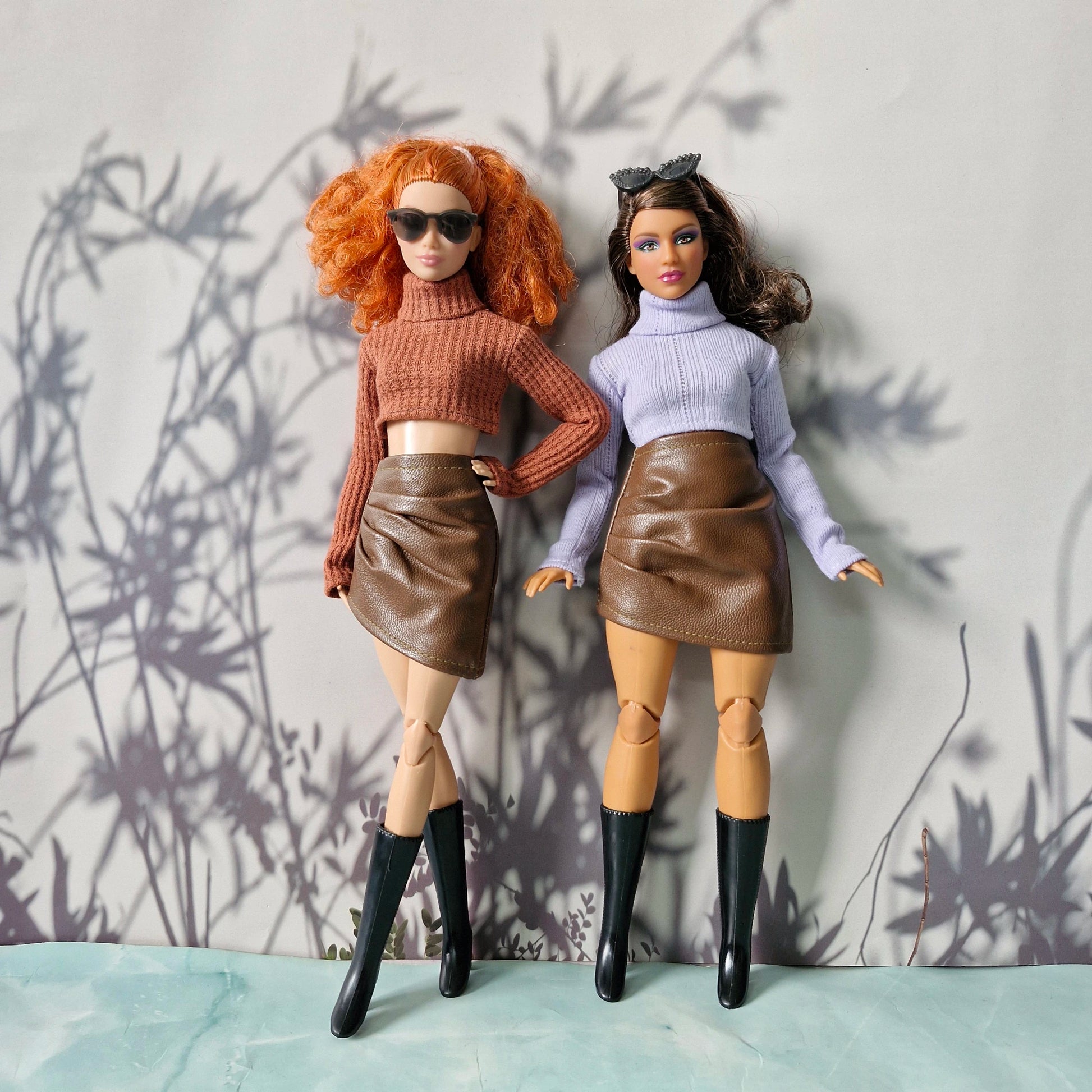Two dolls wearing matching outfits with brown leather skirts and black boots against a neutral background.