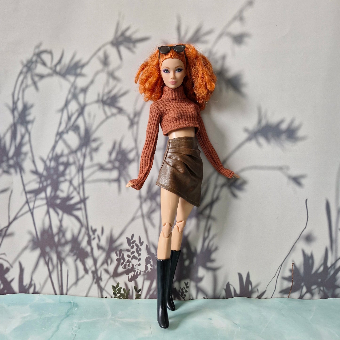Barbie doll with red hair wearing a brown sweater and leather skirt against a textured wall.