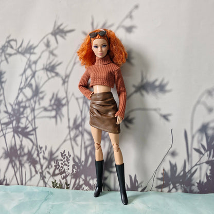Doll wearing a brown sweater and skirt with black boots against a light gray wall with plant shadows.
