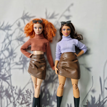 Two dolls wearing matching outfits with brown skirts and turtleneck sweaters against a neutral background.
