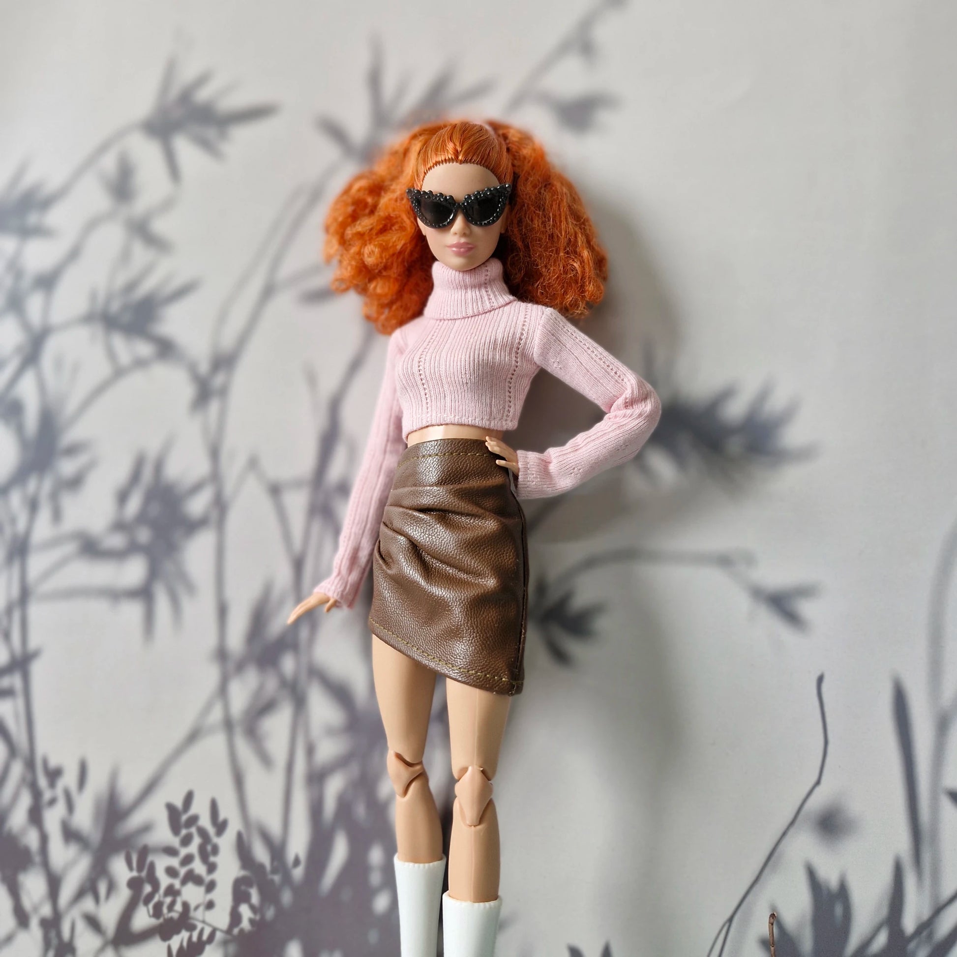 Doll with red hair wearing sunglasses, a pink sweater, and brown skirt against a gray wall with plant shadows.