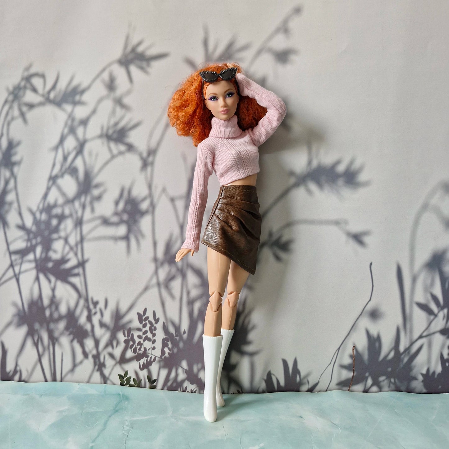 Doll wearing a pink sweater and brown skirt against a wall with plant shadows