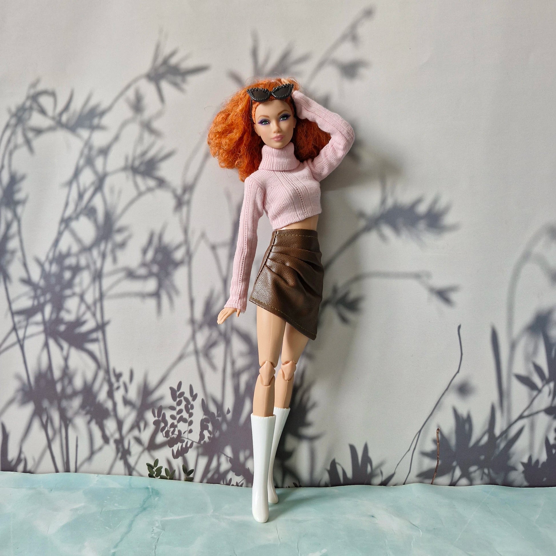Doll wearing a pink sweater and brown skirt against a wall with plant shadows