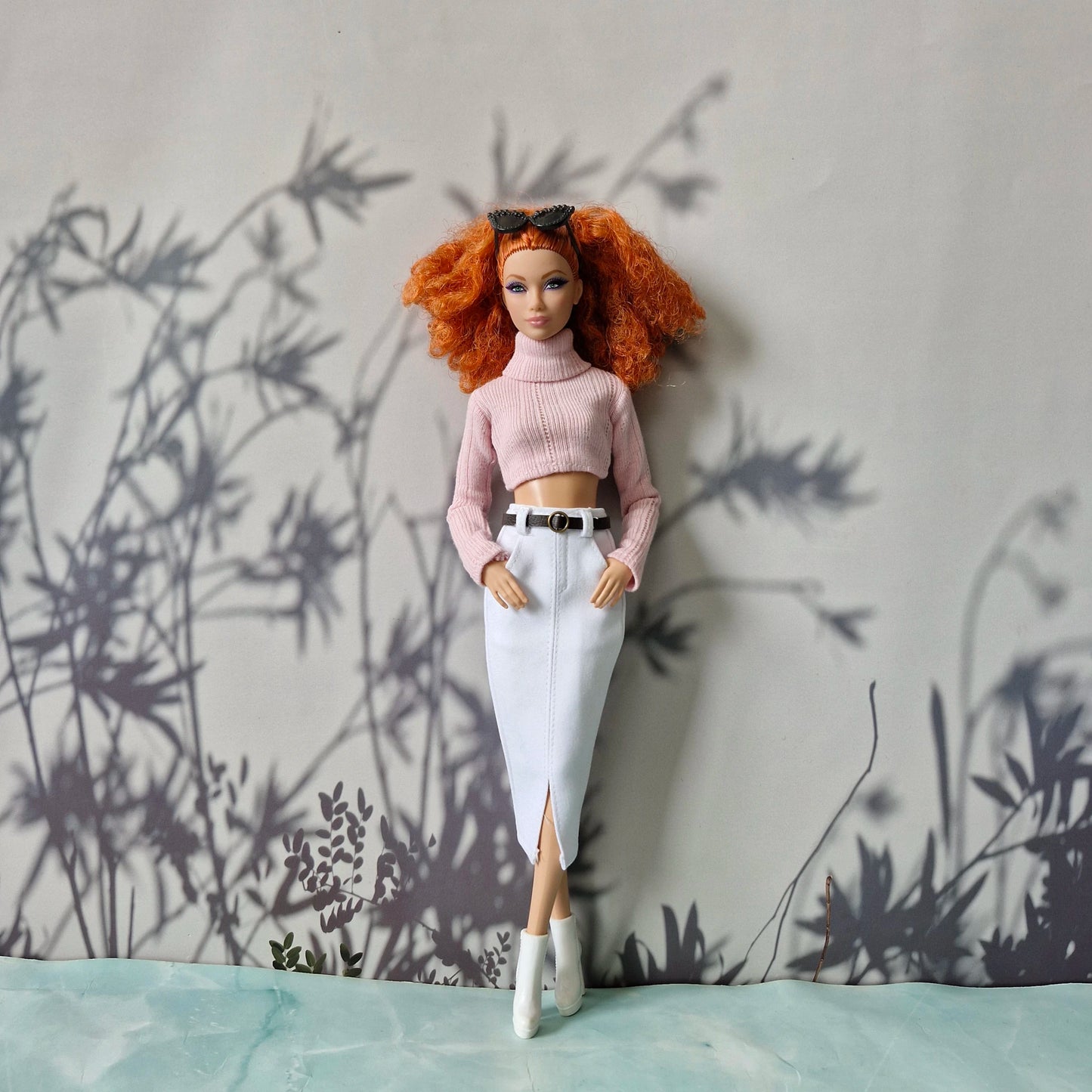 Barbie doll with red hair and a pink outfit standing against a wall with plant shadows.