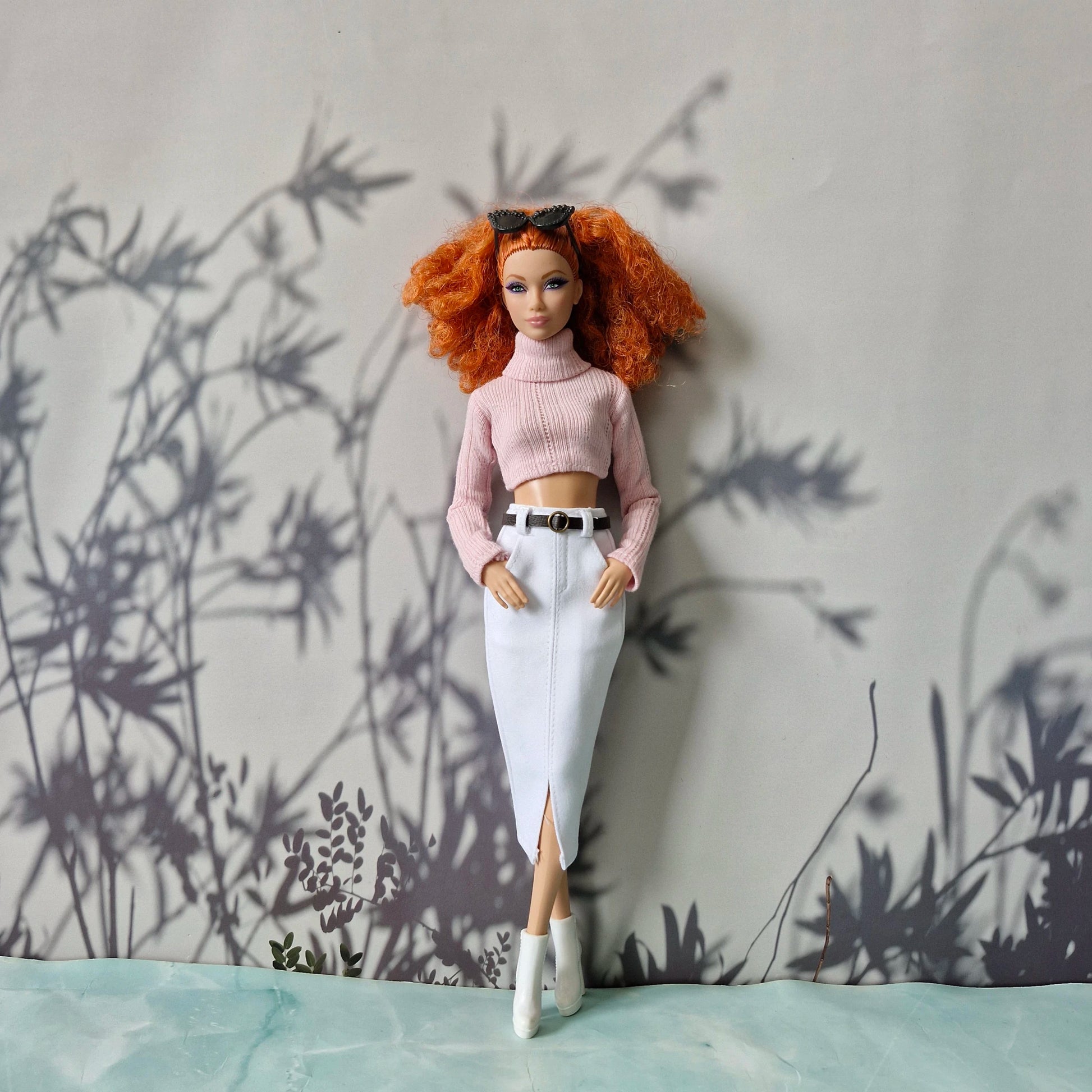 Barbie doll with red hair and a pink outfit standing against a wall with plant shadows.