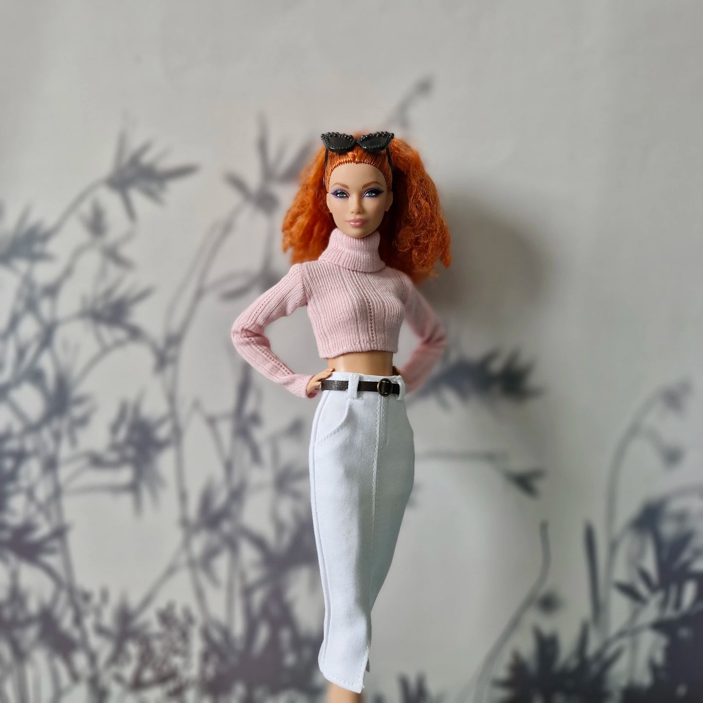 Barbie doll with red hair wearing a pink sweater and white pants against a gray wall.
