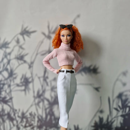 Barbie doll with red hair wearing a pink sweater and white pants against a gray wall.