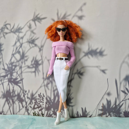 Barbie doll with red hair wearing sunglasses, a pink top, and white pants against a gray wall with plant shadows.