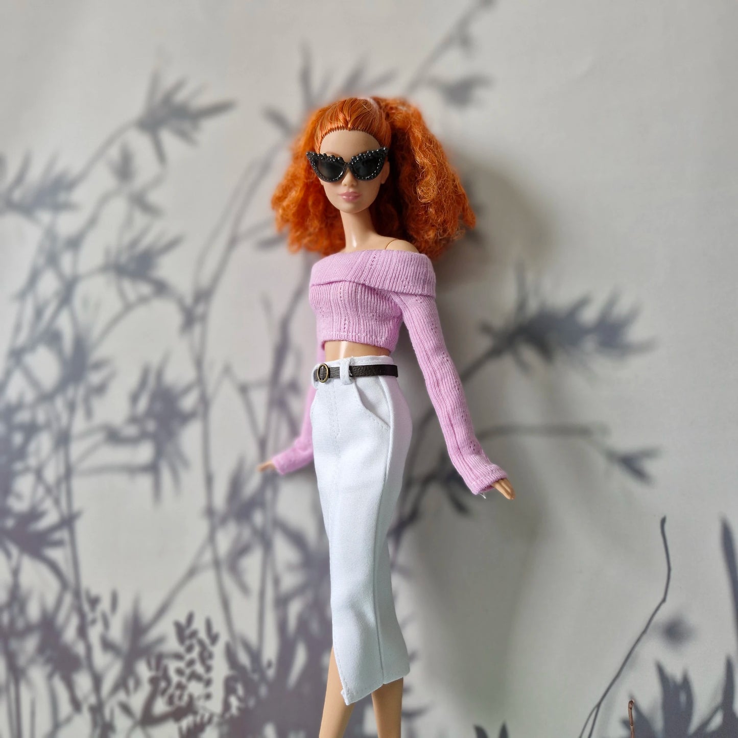 Barbie doll with red hair and sunglasses against a gray wall with plant shadows