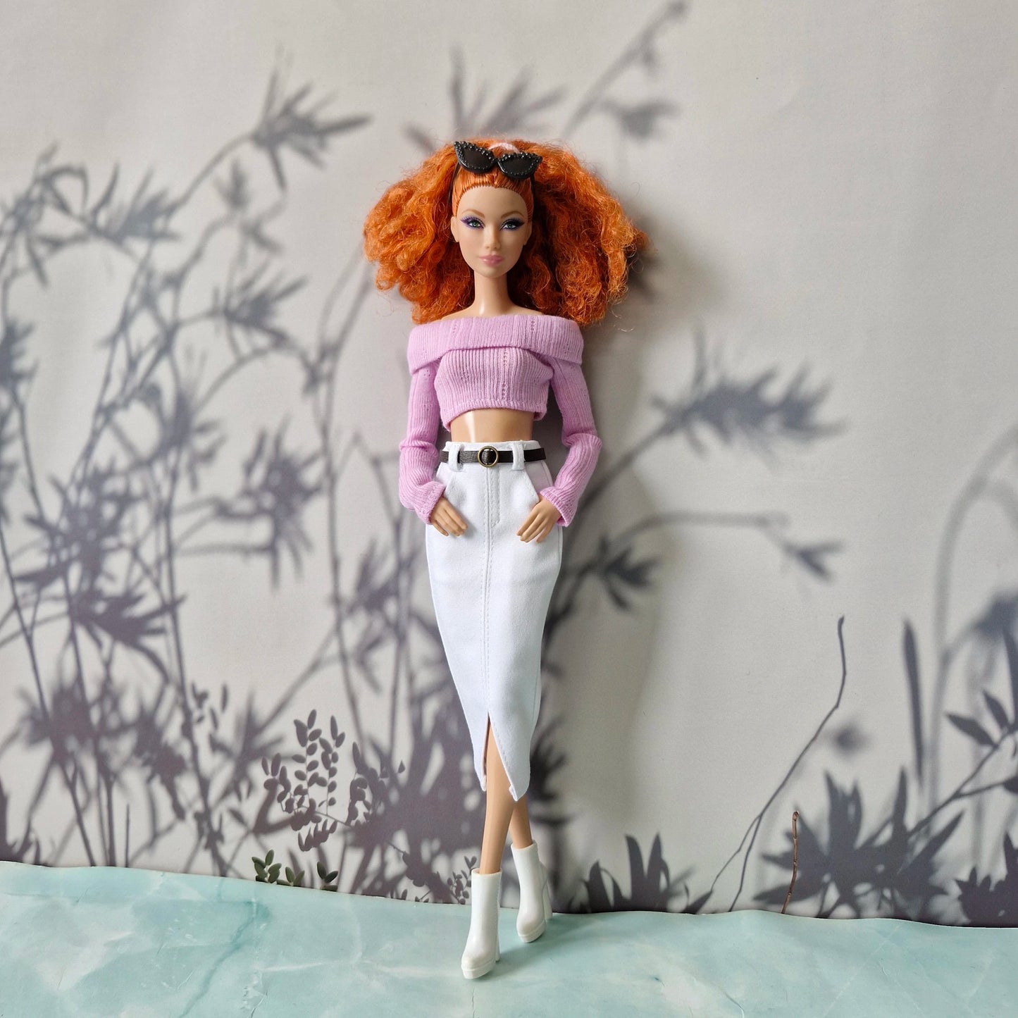 Barbie doll with red hair wearing a pink top and white pants against a wall with plant shadows.