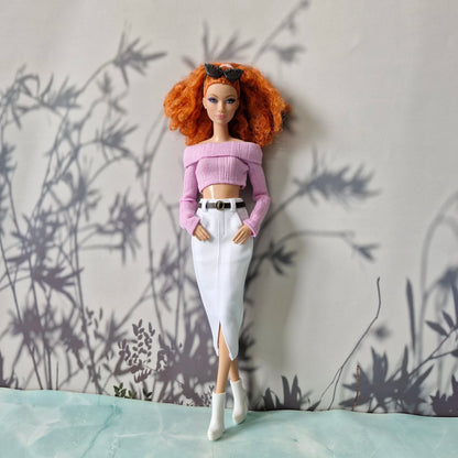 Barbie doll with red hair wearing a pink top and white pants against a wall with plant shadows.