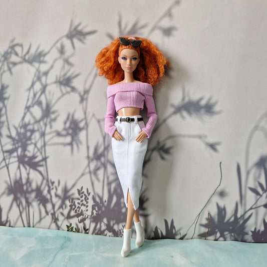 Barbie doll with red hair wearing a pink top and white pants against a wall with plant shadows.