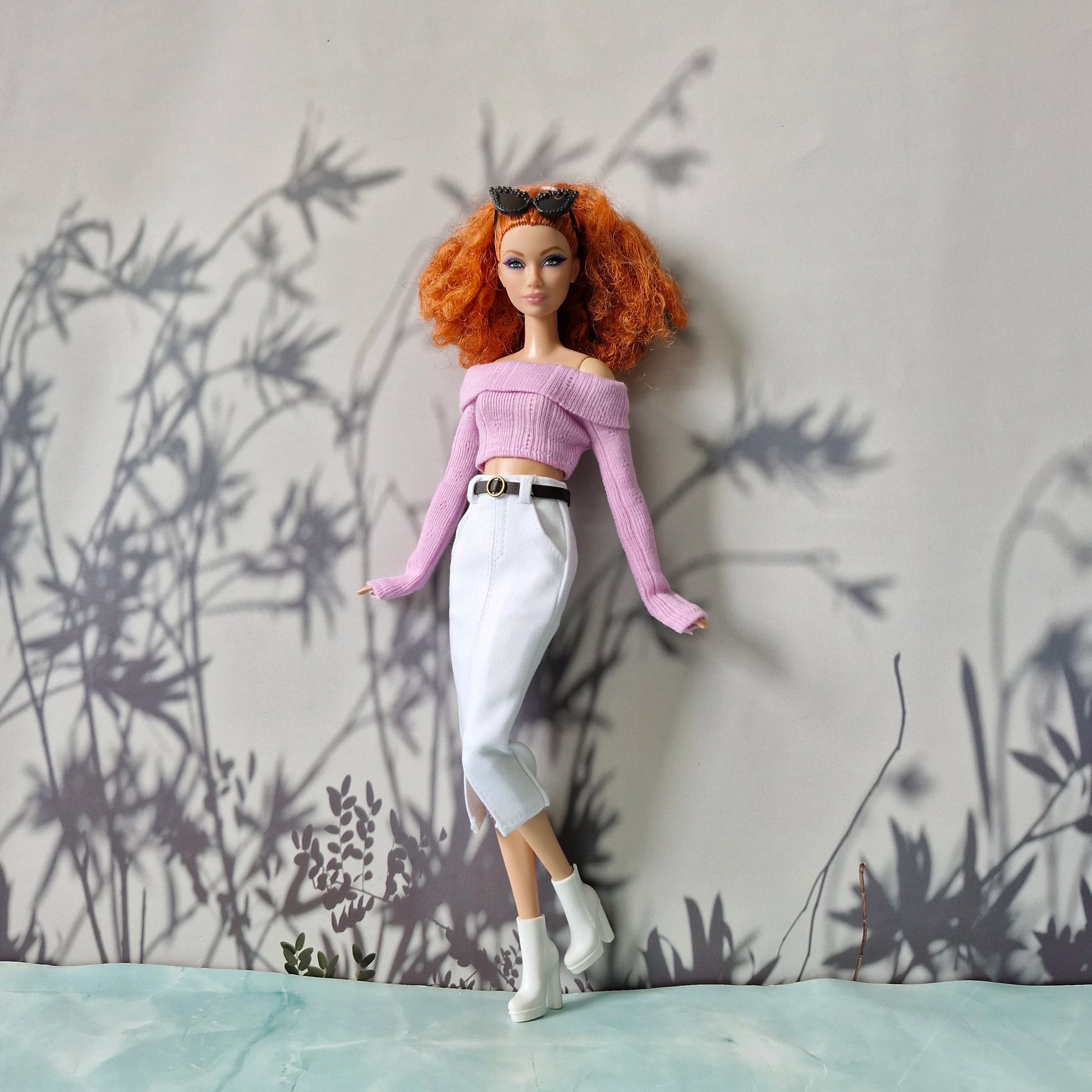 Barbie doll with red hair and a purple top standing against a gray wall with plant shadows.
