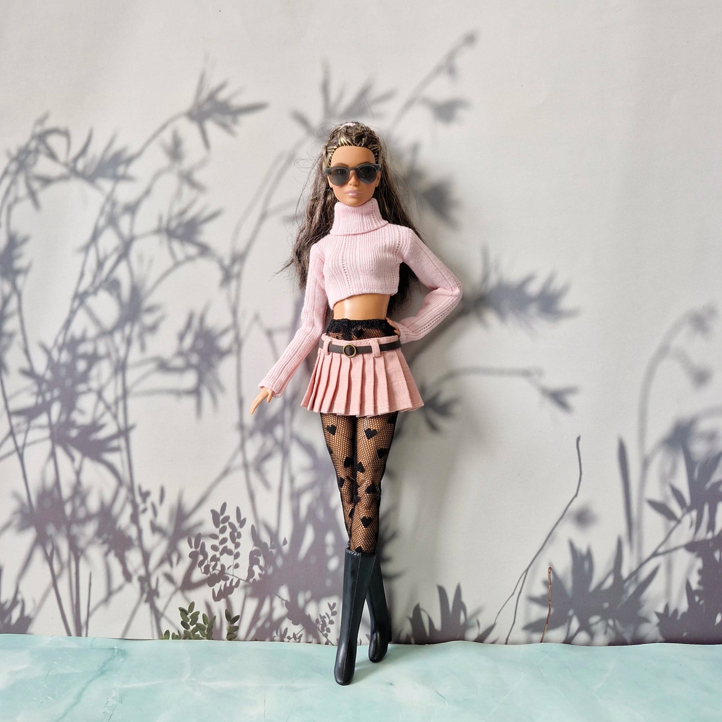 Barbie doll in stylish outfit with pink top, skirt, and black boots against a light gray background with plant shadows.