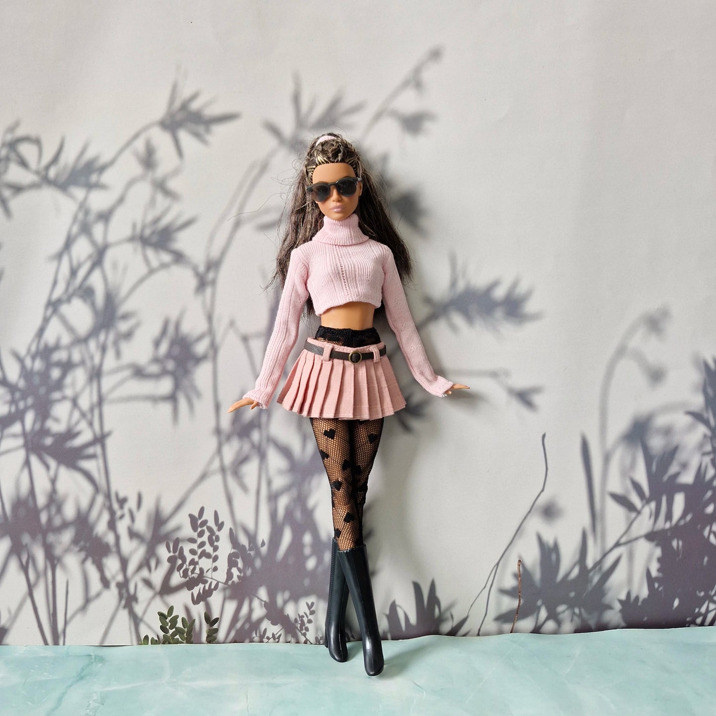 Barbie doll in a stylish outfit with a pink top and skirt, black stockings, and sunglasses, standing against a light gray wall with plant shadows.