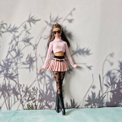 Barbie doll in a stylish outfit with a pink top and skirt, black stockings, and sunglasses, standing against a light gray wall with plant shadows.