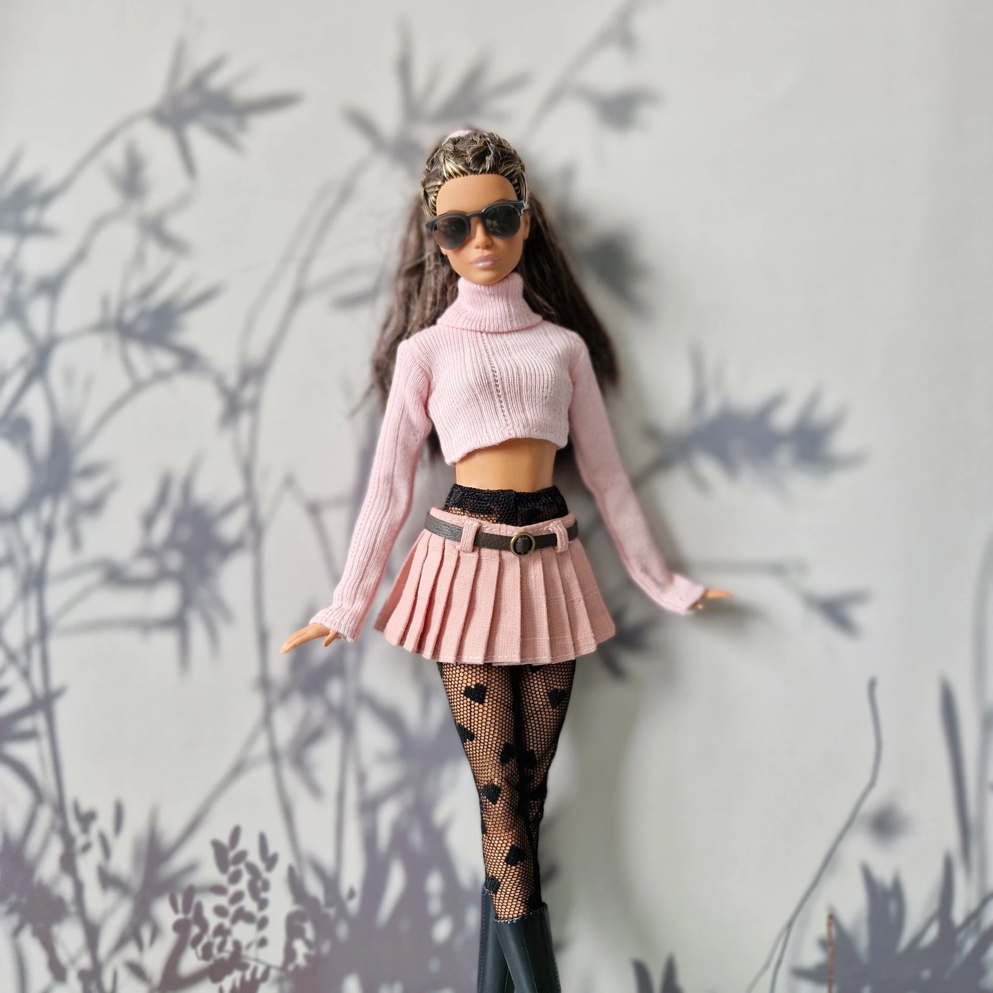Barbie doll wearing a pink outfit with a skirt and turtleneck, standing against a light gray wall with plant shadows.