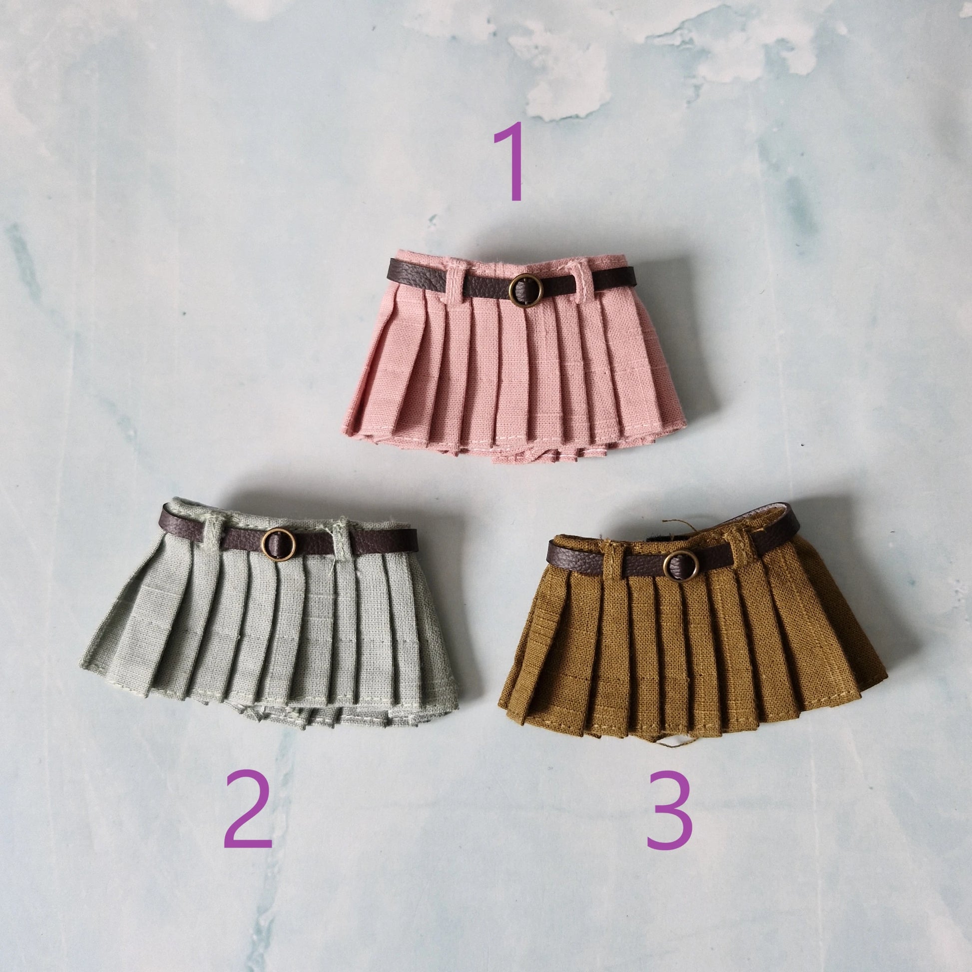 Three pleated skirts in pink, gray, and brown with belts on a textured light background.