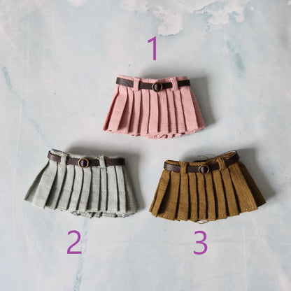 Three pleated skirts in pink, gray, and brown with belts on a textured light background.