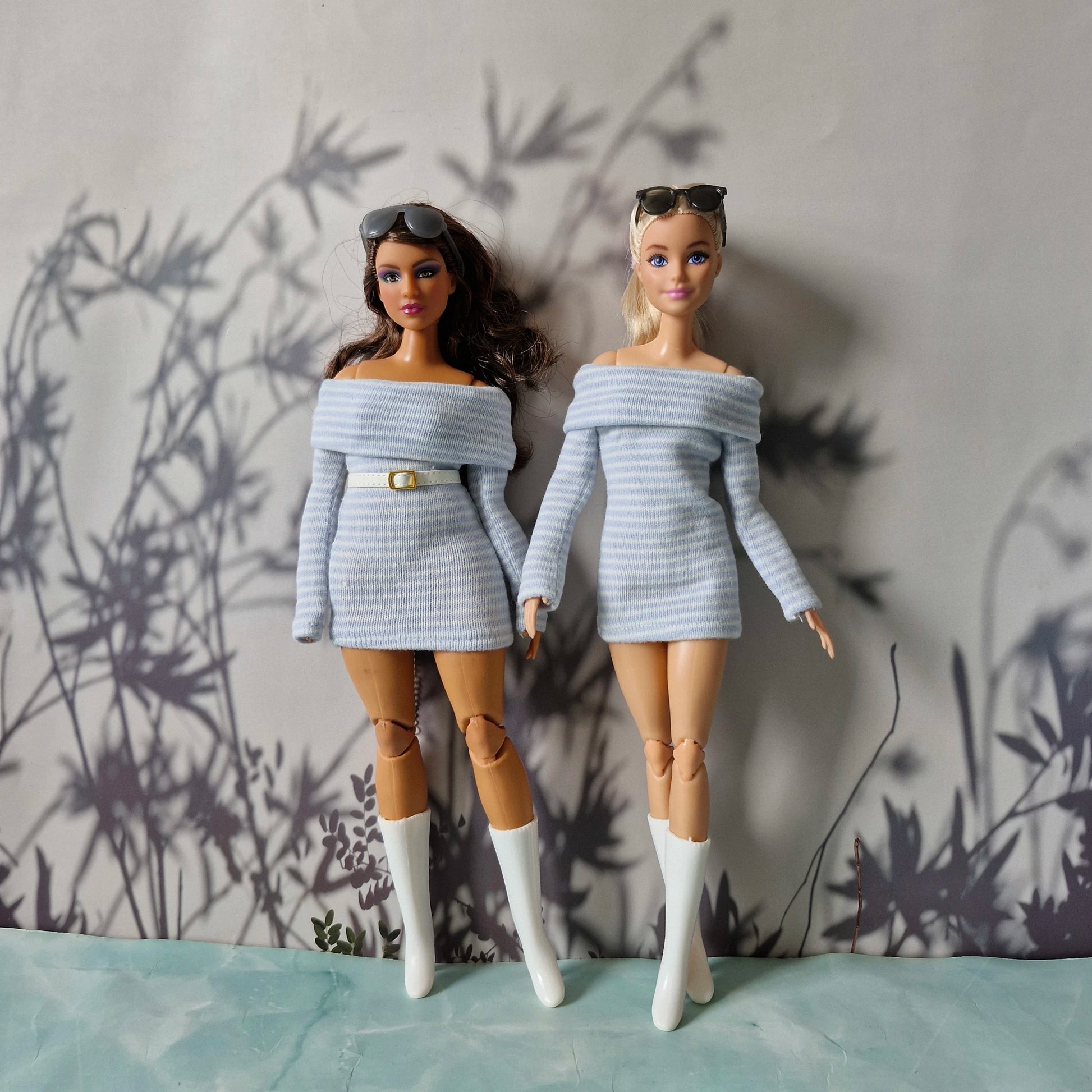Two dolls wearing matching off-shoulder sweaters against a textured wall.