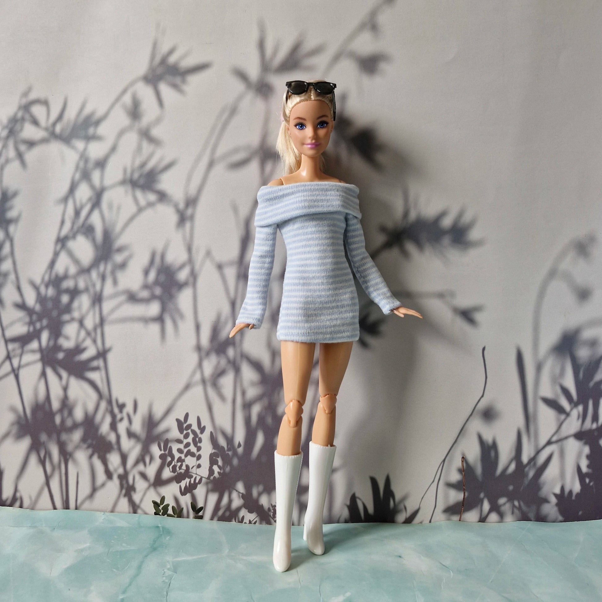 Barbie doll wearing a gray off-shoulder sweater and white boots against a wall with plant shadows.